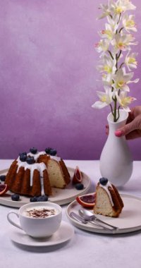 Lezzetli bir pasta, cappuccino ve taze incirle nefis bir sofra kurarken, bir kadın bir vazoda beyaz orkideler düzenleyip sofistike bir atmosfer yaratıyor.