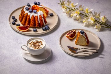 Beyaz kremalı, yaban mersinli ve kanlı portakal dilimli, kapuçino ve orkide ile servis edilen Gugelhupf keki, cezbedici bir masa hazırlıyor.