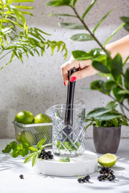 Woman using muddler to crush mint leaves, blackcurrants, and lime in a mixing glass, preparing a refreshing cocktail clipart