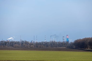 Yeşil bir tarla ve ormanın arkasındaki sanayi bölgesinden yükselen duman çevresel etkiyi vurguluyor.