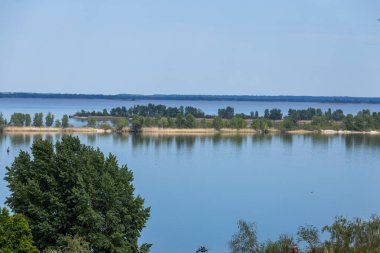 Sakin göl, açık mavi gökyüzünün altındaki ağaçlar ve bitki örtüsünü yansıtıyor. Huzurlu ve huzurlu bir doğal manzara yaratıyor.