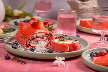 Krem peynirle dolu dilimlenmiş pembe çilekli kek rulosu, taze çilek, yaban mersini ve pembe masa örtüsü üzerine beyaz çiçekler.