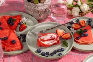 Krem peynir ve çilekle dolu pembe rulo krep, kırmızı gözlemelerle eşleştirilmiş, canlı yeşil tabaklarda taze böğürtlen