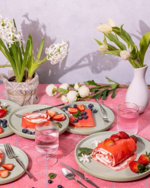 Çilek, yaban mersini ve nane şekeriyle süslenmiş pembe krep rulolarının yanında pembe kiremitli bir masada su ve çiçekler.