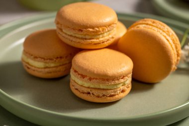 Kremalı canlı turuncu badem kurabiyeleri yeşil bir tabakta oturur ve görsel olarak çekici bir kontrastlık yaratır.