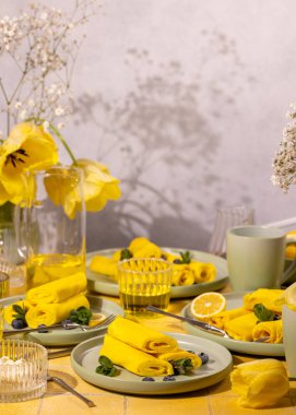 Yaban mersini, nane ve limon dilimleri ile servis edilen lezzetli altın krepler, canlı sarı bir kahvaltı ortamı yaratıyor.