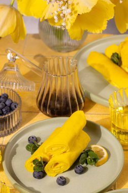 Sarı laleli sarı bir masada yabanmersini, nane ve limon ile servis edilen yuvarlak sarı krepler kahve eşliğinde.