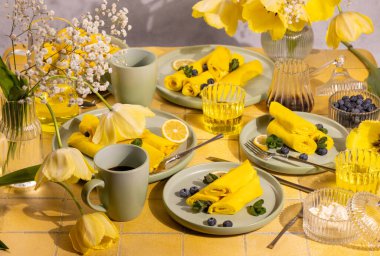 Yaban mersinli ve nane şekerli leziz limonlu krepler sarı lalelerle süslenmiş bir masada, canlı ve iştah açıcı bir kahvaltı sahnesi yaratıyor.