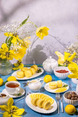 Sarı limonlu badem kurabiyeleri, sıcak çay ve taze limonla servis edilir mavi bir masada, sarı lale ve goji üzümleriyle süslenir.