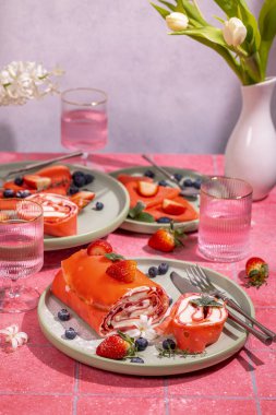 Krem peynirli kırmızı kadife kek. Taze çilek ve yaban mersiniyle süslenmiş. Pembe masa ortamında servis ediliyor.