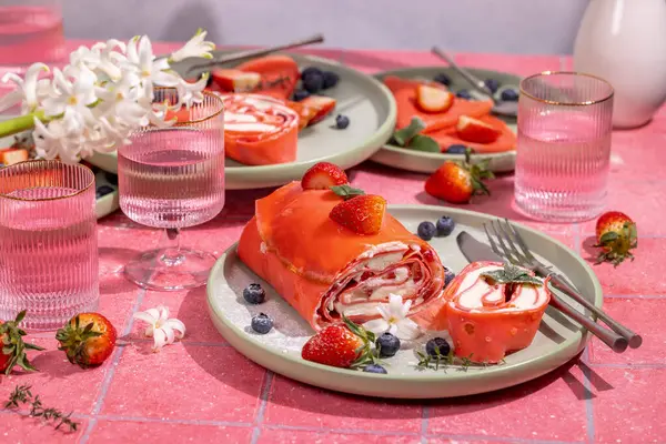 Lezzetli kırmızı çilekli krepler taze çilekler, yabanmersinleri ve tazeleyici suyla eşleştirilmiş narin beyaz çiçeklerle servis edilir.