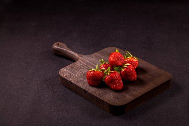 Olgun, canlı çilekler koyu, ahşap bir kesme tahtasının üzerinde oturur, basit ama çekici bir mutfak sahnesi yaratır.