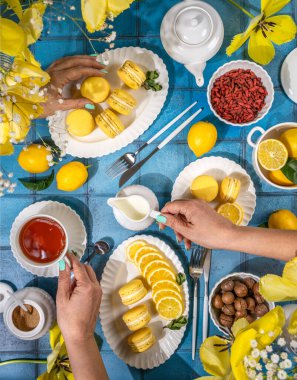 İki kadın limonlu badem kurabiyesi, çay, dilimlenmiş limon ve sarı lalelerle süslenmiş mavi bir masada çeşitli ikramlarla çay saatlerini paylaşıyor.
