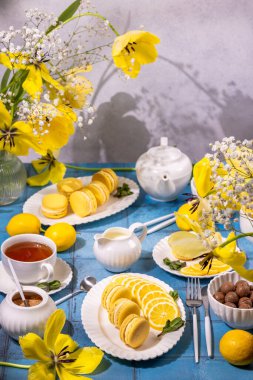 Sarı laleler ve çingenelerle süslenmiş mavi bir masada çay ve limon dilimleri ile servis edilen lezzetli limonlu kurabiyeler.