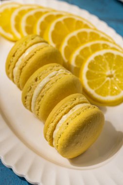 Canlı sarı limonlu badem kurabiyeleri, zarif beyaz bir tabakta taze kesilmiş limon dilimlerinin yanında servis edilir ve görsel olarak çekici bir tatlı sunumu oluşturur.