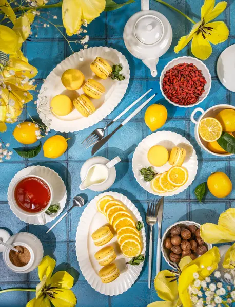 Limonlu badem kurabiyesi, dilimlenmiş limon, goji üzümü, çay ve sarı lalelerin yer aldığı bahar kahvaltı masası. Mavi fayanslı bir yüzeye dizilmiş.