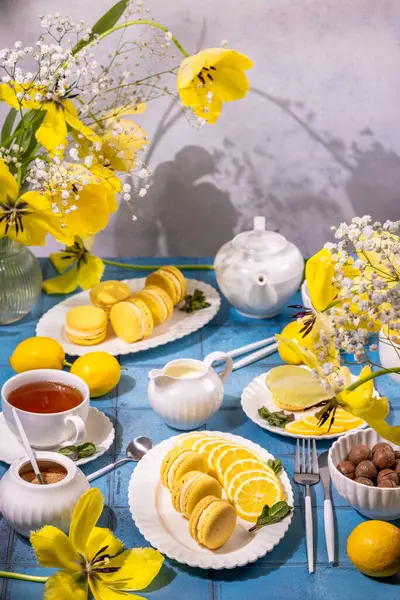 Sarı laleler ve çingenelerle süslenmiş mavi bir masada çay ve limon dilimleri ile servis edilen lezzetli limonlu kurabiyeler.
