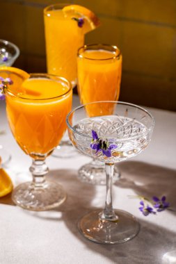 İçlerinde taze portakal suyu kokteylleri bulunan kristal bardaklar yenilebilir çiçekler ve portakal dilimleriyle süslenerek sofistike ve canlı bir içecek sunumu oluşturulur.