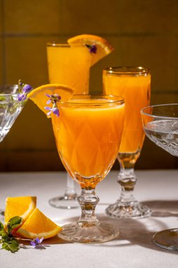 Canlı portakal suyu kokteylleriyle dolu kristal bardaklar, yenilebilir çiçekler ve portakal dilimleriyle süslenmiş, canlandırıcı ve görsel olarak çekici bir sahne yaratır.