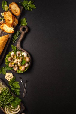 Sarımsaklı salyangoz, maydanoz, biberiye, limon, baget ve siyah arka planda servis edilen özel çatallar.