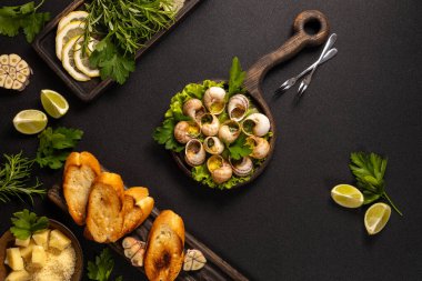 Sebzeli gurme salyangozlar, sarımsaklı tereyağı ve kızarmış ekmek dilimleri siyah masa ortamında leziz mutfak sahneleri yaratıyor.