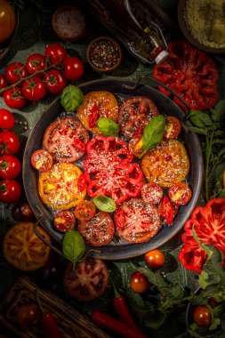 Farklı renkte ve boyutlarda dilimlenmiş organik domatesler, rendelenmiş peynir, fesleğen ve kırmızı biberle süslenmiş koyu bir kırsal tabakta, canlı bir mutfak sahnesi oluşturur.