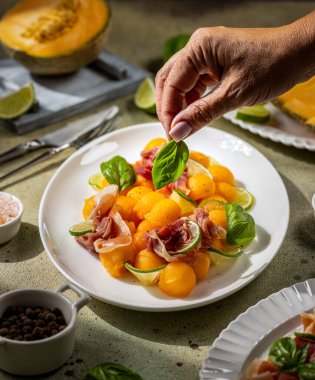 Şefin taze fesleğen yaprağı jambonlu kavun salatası, limon dilimleri ve bir tutam karabiber ekleyip ferahlatıcı ve lezzetli bir yaz yemeği hazırlaması.