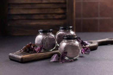 Mor renkli tuz içeren üç küçük cam kavanoz taze mor fesleğenle süslenmiş ve kırsal bir ahşap kesme tahtasına yerleştirilmiş.