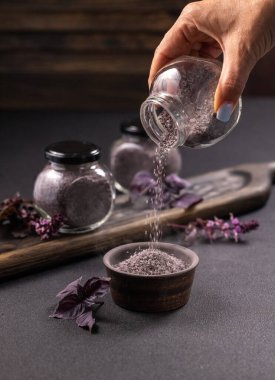 Cam bir baharat kavanozundan taze fesleğen ve koyu ahşap bir tahtanın üzerine baharat dolu kavanozlar içeren küçük bir seramik kaseye mor mineral kristal tuz dökerek.