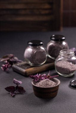 Mor fesleğen yaprakları ve çiçek açan filizler bir kase bitkisel tuzu süslüyor, kırsal bir ahşap tahtada cam kavanozlarda daha fazla tuz depolanarak karanlık ve durgun bir yaşam kompozisyonu oluşturuyor.