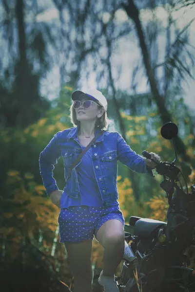 站在自然背景下的小摩托车旁边的漂亮女人 免版税图库照片
