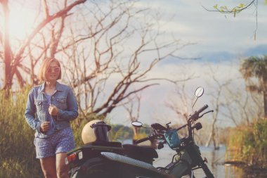 Güzel dişlek kadın mutluluktan gülümsüyor orman tarlasında küçük bir dayanıklı motosikletin yanında duruyor.