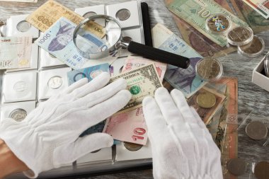 Ellerinde farklı eski koleksiyoncu paraları ve büyüteçle banknotlar var.