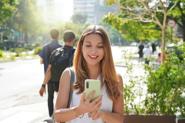 Young business woman walking and reading text on mobile phone in city street on summer time with blurred background. Sustainable city lifestyle people technology.