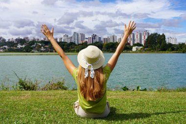 Young woman with hat raising arms sitting on grass in Barigui Park, Curitiba, Brazil clipart