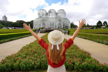 Tourism in Curitiba, Brazil. Back view of stylish traveler woman with raising arms in  botanical garden of Curitiba, Parana, Brazil. clipart