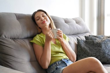 Kapalı gözlü, bitkisel çay içen, evdeki rahat kanepesinde rahatlamış genç bir kadının portresi.