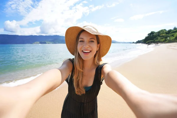 Laughing girl takes self portrait on empty tropical island in her summer vacation. Selfie of smiling tourist woman on Ilhabela Island beach, Brazil.