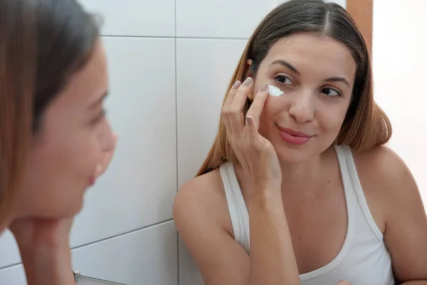 Beauty routine at home. Portrait of beautiful girl applying cream under eye with finger. Dark circles and anti-wrinkle concept.