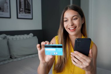 Çevrimiçi ödeme için kredi kartı numarasını okuyan ve akıllı telefonunu evde tutan gülen kızın portresi.