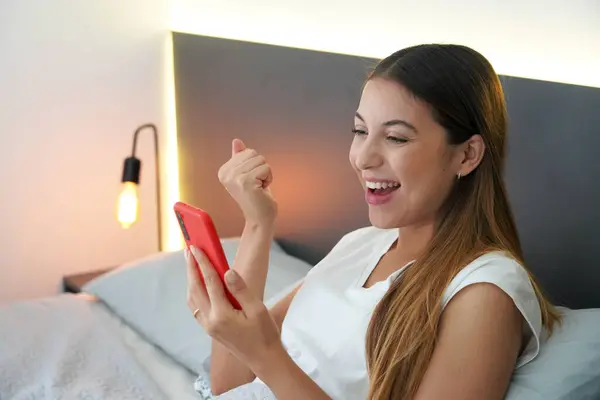 Excited young woman in bed looking at her smartphone with amazed surprised face celebrating victory or good news