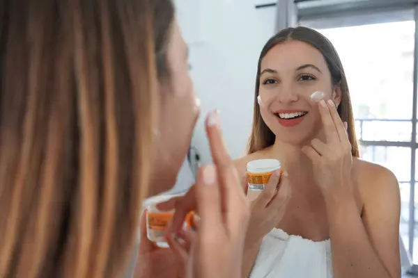 Beautiful young woman applying cream on her cheeks. Face moisturizing nourishing invigorating treatments. Enjoying relaxing time of skin care on mirror at home.