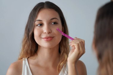 Yüz kılları alınıyor. Dermplaning. Güzel bir kızın evde jiletle yüzünü tıraş edişinin yakın çekimi. Aynada saçlarını kesmek için jilet kullanan hoş bir genç kadın..
