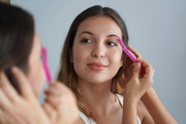 Dermplaning. Evdeki aynada jiletle kaşlarını tıraş eden güzel bir kızın yakın çekimi. Yüz kılları alınıyor..