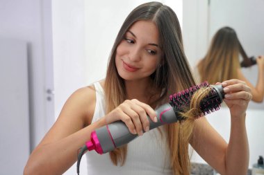 Hot air hair brush. Portrait of young woman using round brush hair dryer to style hair in an easy way at home. Girl using electric blowout brush hair dryer. clipart