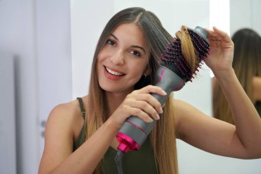 Hot air hair brush. Young woman using round brush hair dryer to style hair at home. Teenager girl using electric blowout brush hair dryer. Looking at camera. clipart