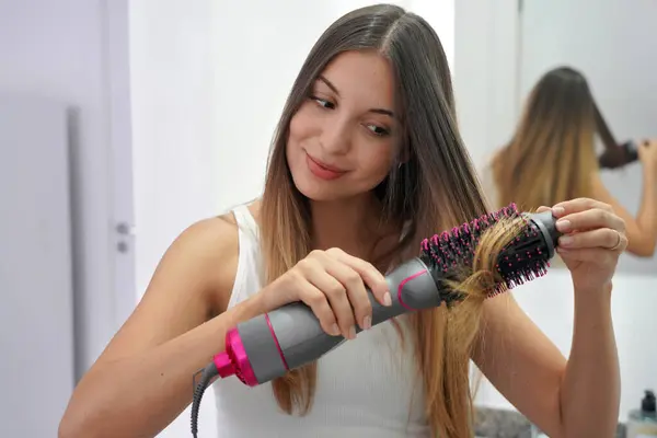 Hot air hair brush. Portrait of young woman using round brush hair dryer to style hair in an easy way at home. Girl using electric blowout brush hair dryer.