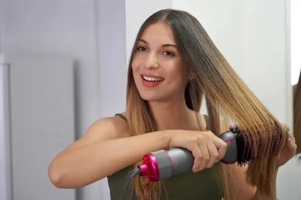 Hot air hair brush. Young woman using round brush hair dryer to style hair in an easy way at home. Teenager using electric blowout brush hair dryer.