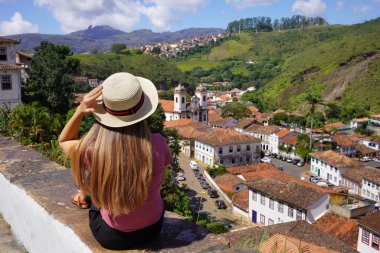 Şapkalı turist kız duvarda oturmuş tarihi Ouro Preto, Minas Gerais, Brezilya 'nın panoramik manzarasına bakıyor.