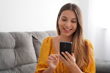 Latin kökenli çekici genç bir kadın oturma odasında akıllı telefonunu kullanıyor.